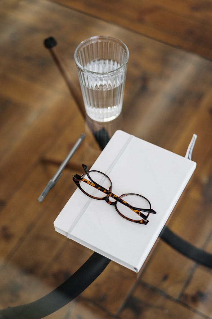 A stylish and minimalistic top view of a workspace with glasses, a notebook, and a glass of water on a wooden table.